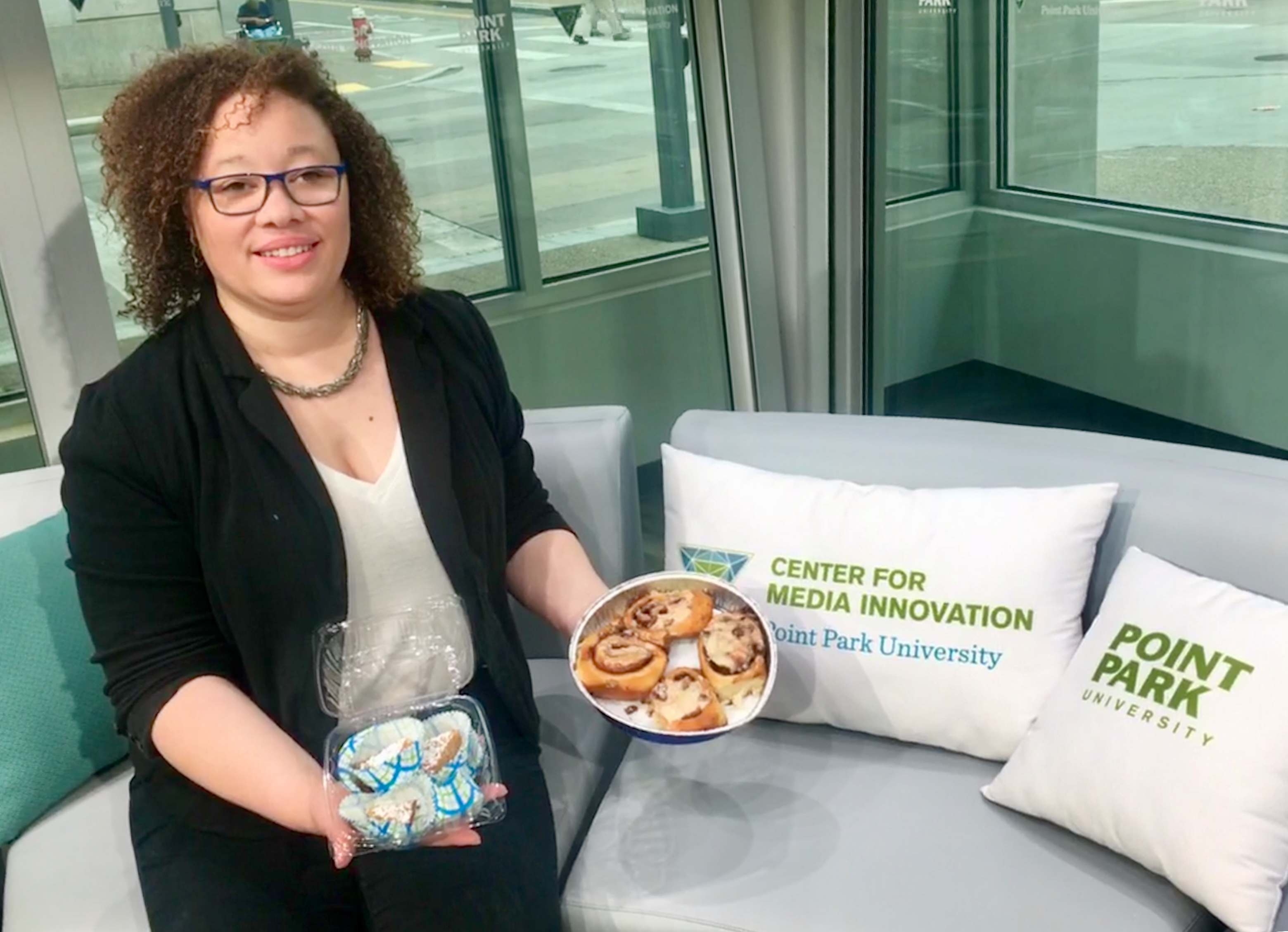 A woman in a white blouse and black blazer has homemade desserts in her hand, including chess pie bites and glazed cinnamon buns. She's on the interview couch in the TV studio at the Center for Media innovation at Point Park University.